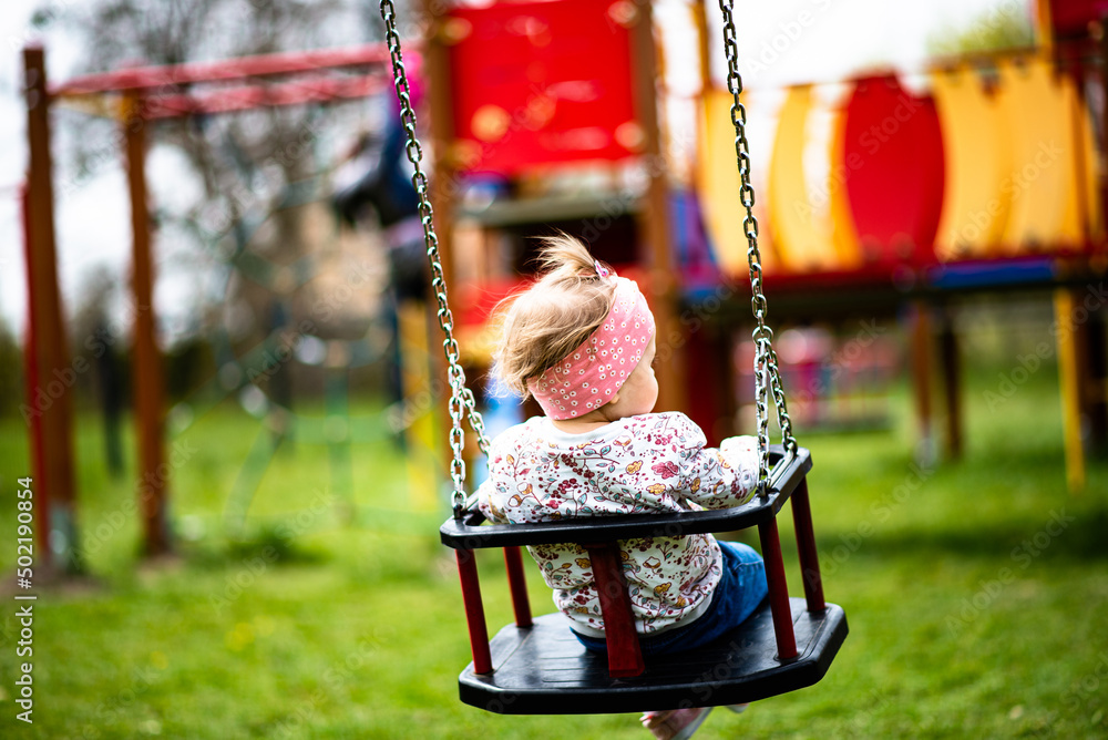 baby girl is swinging on a swing at blurred playground background - obrazy, fototapety, plakaty 