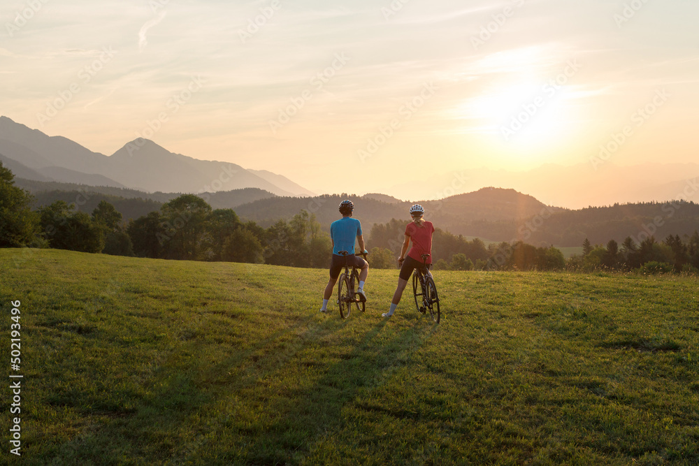 Professional cyclist couple taking a break on a green meadow and watching amazing mountain sunset, aerial shot.