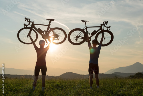 Two cyclers silhouettes holding road bicycles high above heads, in winner pose during sunset over the mountain landscape