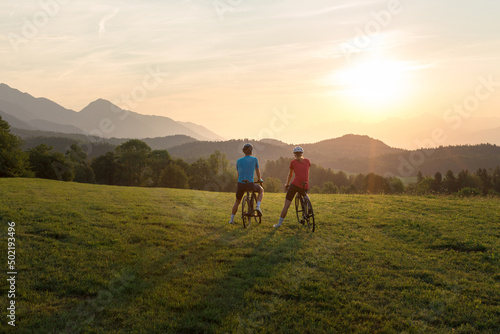 Professional cyclist couple taking a break on a green meadow and watching amazing mountain sunset, aerial shot.