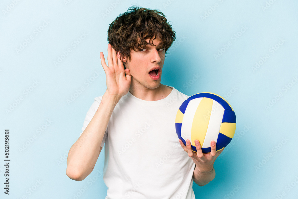 Young caucasian man playing volleyball isolated on blue background trying to listening a gossip.