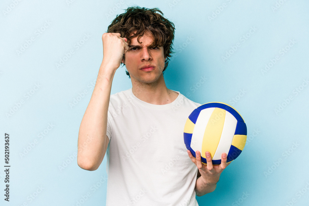 Young caucasian man playing volleyball isolated on blue background showing fist to camera, aggressive facial expression.