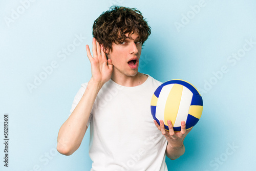 Young caucasian man playing volleyball isolated on blue background trying to listening a gossip.