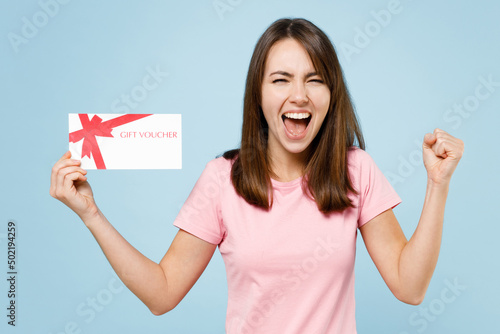 Young excited happy caucasian woman 20s in pink t-shirt hold gift certificate coupon voucher card for store do winner gesture isolated on pastel plain light blue background. People lifestyle concept.