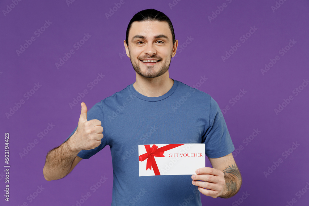 Young smiling happy man 20s wearing basic blue t-shirt hold gift certificate coupon voucher card for store show thumb up isolated on plain purple background studio portrait. People lifestyle concept. Young smiling happy man 20s wearing basic blue t-shirt hold gift certificate coupon voucher card for store show thumb up isolated on plain purple background studio portrait. People lifestyle concept.