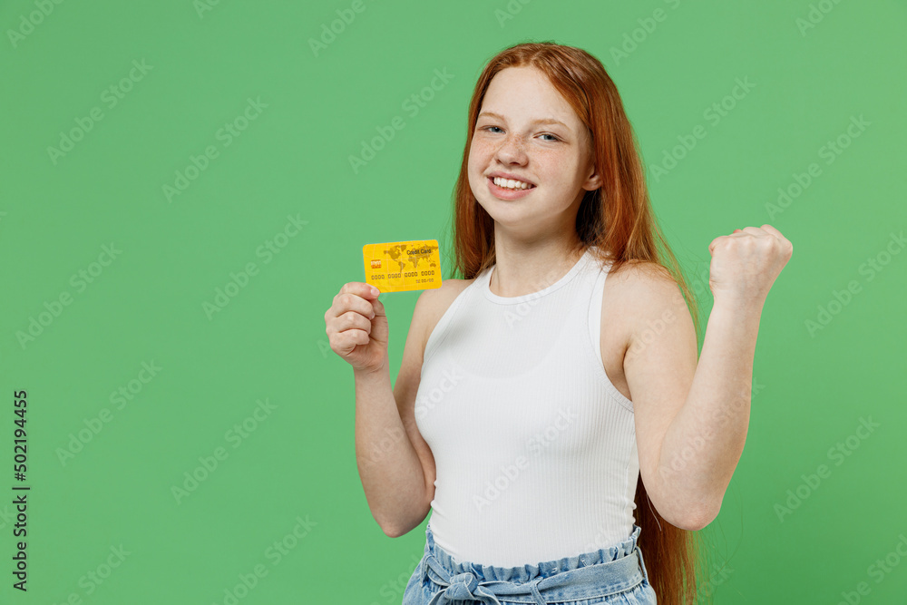 Little redhead kid smiling happy fun girl 12-13 years old wear white tank shirt hold in hand credit bank card do winner gesture isolated on plain green background studio. Childhood lifestyle concept. Little redhead kid smiling happy fun girl 12-13 years old wear white tank shirt hold in hand credit bank card do winner gesture isolated on plain green background studio. Childhood lifestyle concept.