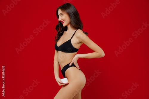 Profile young sexy woman 20s with perfect fit body wear black underwear applying moisturizer, body anti-cellulite cream from container isolated on plain red background. People female beauty concept