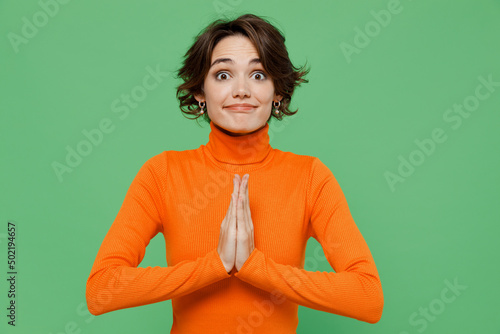 Young pleaded woman 20s in orange turtleneck hold hands folded in prayer gesture, beg about something isolated on plain pastel light green color background studio portrait People lifestyle concept