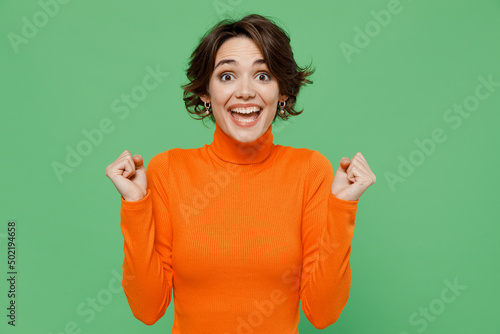 Fotobehang Young smiling woman 20s wear orange turtleneck doing winner gesture celebrate clenching fists say yes isolated on plain pastel light green color background studio portrait
