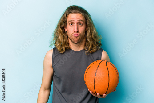 Young caucasian man playing basketball isolated on blue background shrugs shoulders and open eyes confused.