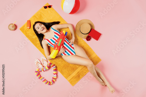 Top view full body young woman of Asian ethnicity in striped swimsuit lies on towel hotel pool hold shoot from water gun isolated on plain pink background. Summer vacation sea rest sun tan concept.