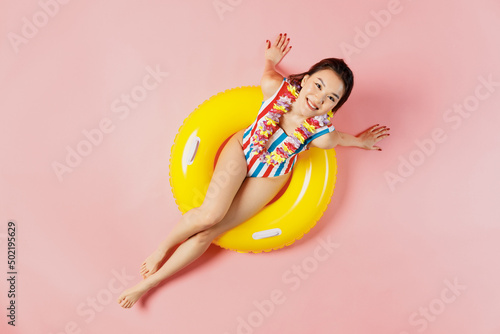 Top view full body smiling young woman of Asian ethnicity in striped swimsuit lies on inflatable rubber ring in pool isolated on plain pastel pink background. Summer vacation sea rest sun tan concept.