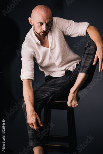 A bald guy with a beautiful beard in a white shirt and plaid pants on a bar stool on a black background