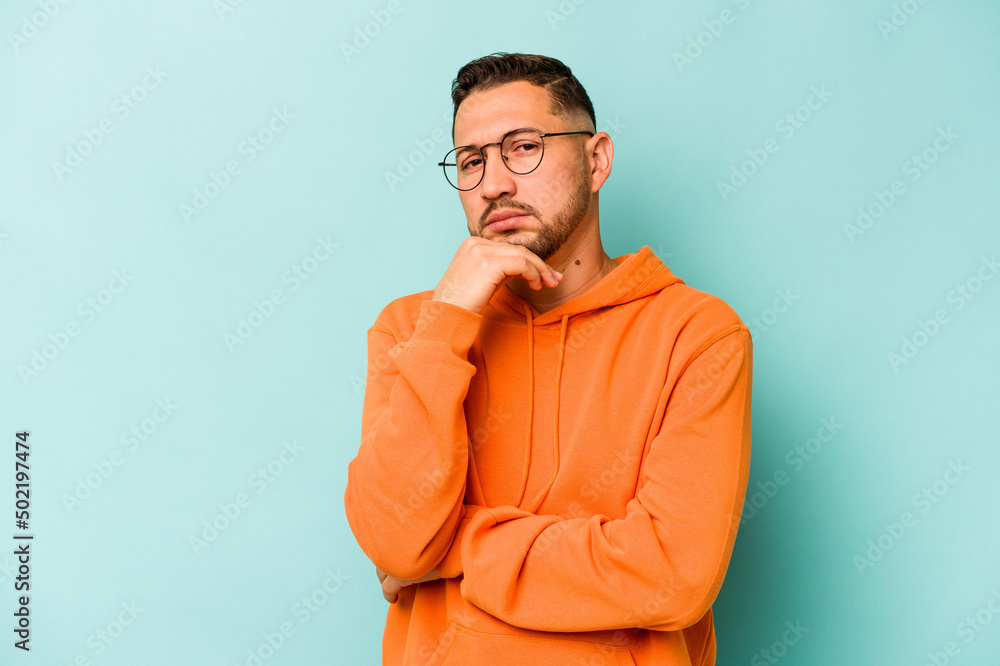 Young hispanic man isolated on blue background suspicious, uncertain, examining you. Young hispanic man isolated on blue background suspicious, uncertain, examining you.