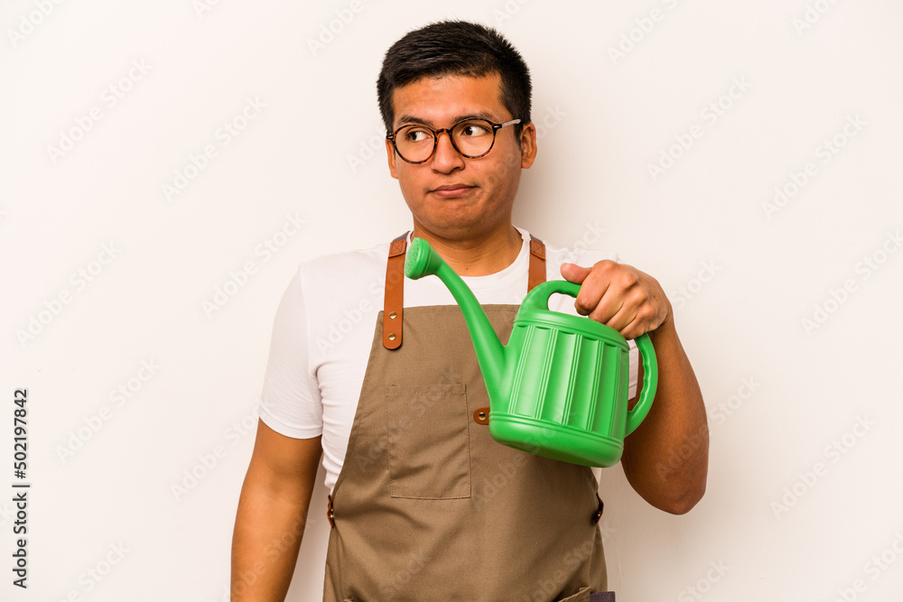 Young gardener hispanic man holding a watering can isolated on white background confused, feels doubtful and unsure. Young gardener hispanic man holding a watering can isolated on white background confused, feels doubtful and unsure.
