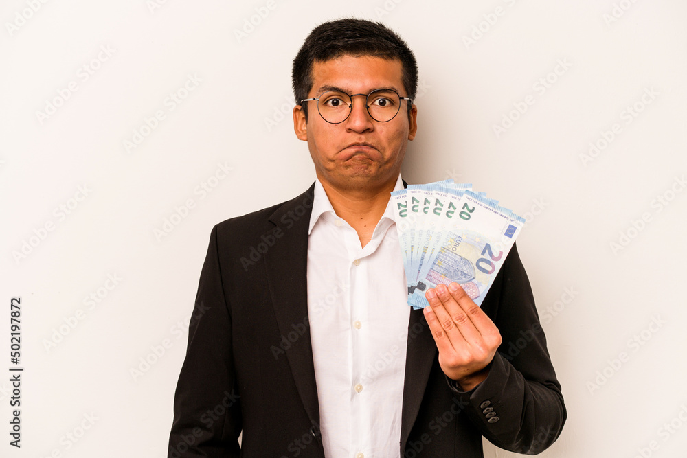 Young hispanic business man holding banknotes isolated on white background shrugs shoulders and open eyes confused. Young hispanic business man holding banknotes isolated on white background shrugs shoulders and open eyes confused.