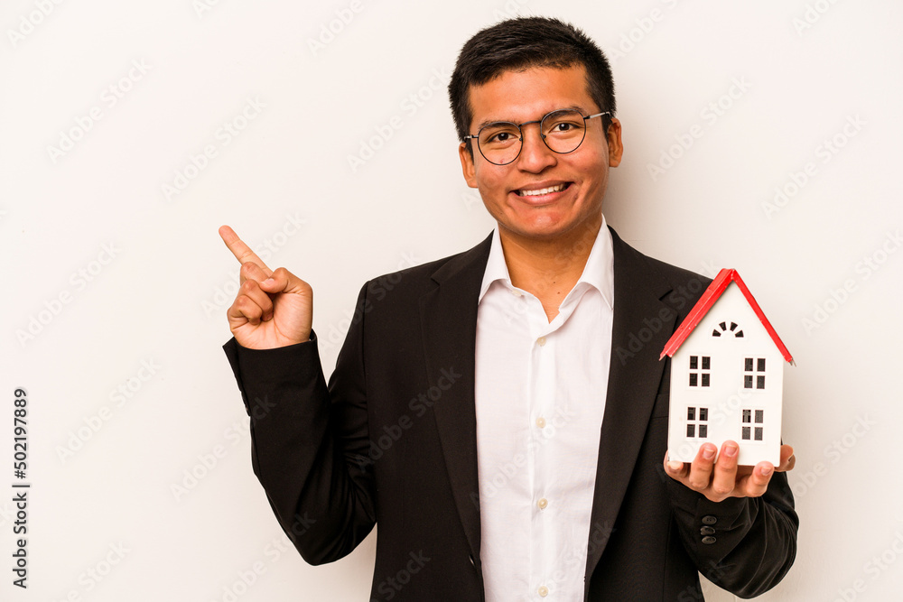 Young business hispanic man holding a little house isolated on white background smiling and pointing aside, showing something at blank space. Young business hispanic man holding a little house isolated on white background smiling and pointing aside, showing something at blank space.
