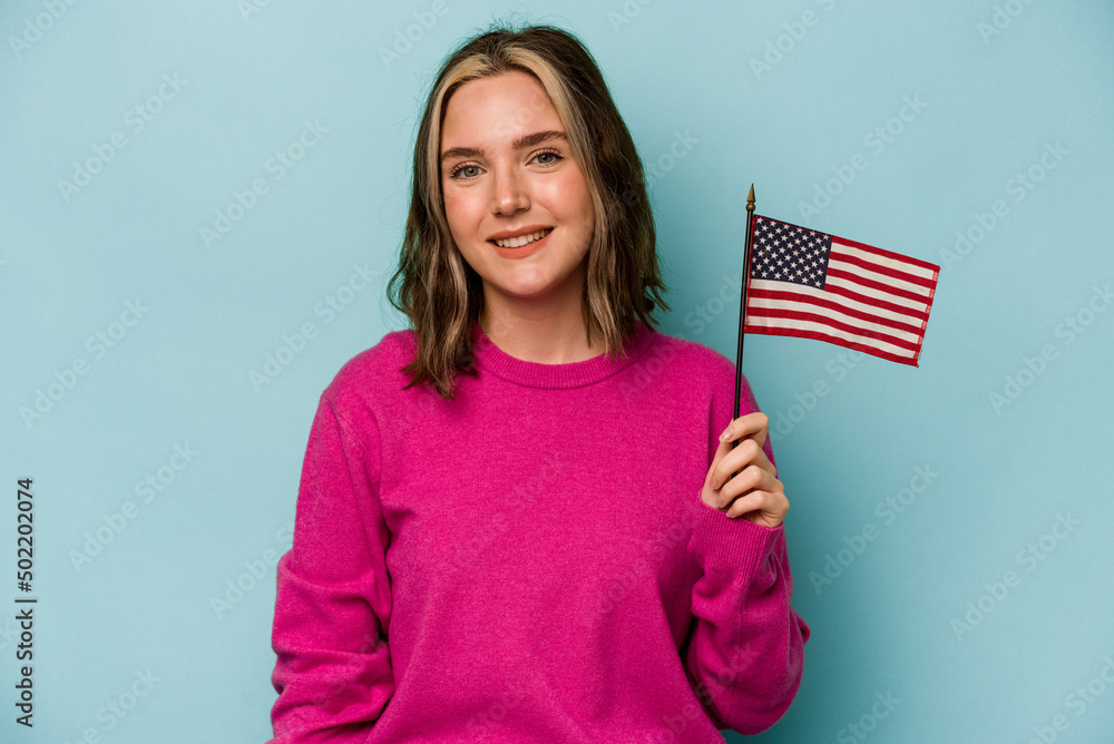 Young caucasian woman holding a American flag isolated on blue background happy, smiling and cheerful. Young caucasian woman holding a American flag isolated on blue background happy, smiling and cheerful.