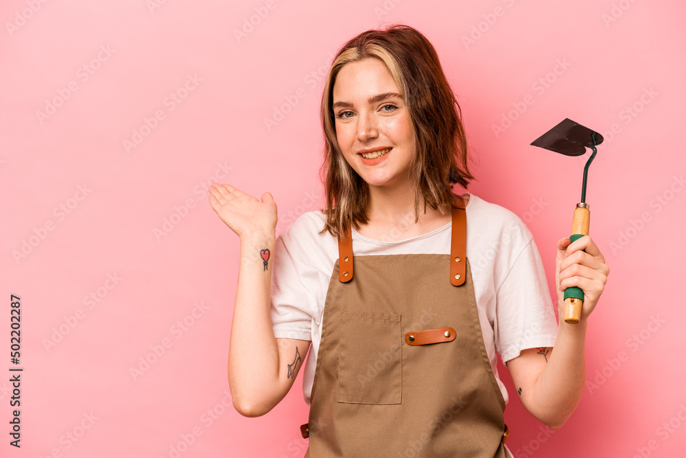 Young gardener woman holding gardening shovel isolated on pink background showing a copy space on a palm and holding another hand on waist. Young gardener woman holding gardening shovel isolated on pink background showing a copy space on a palm and holding another hand on waist.