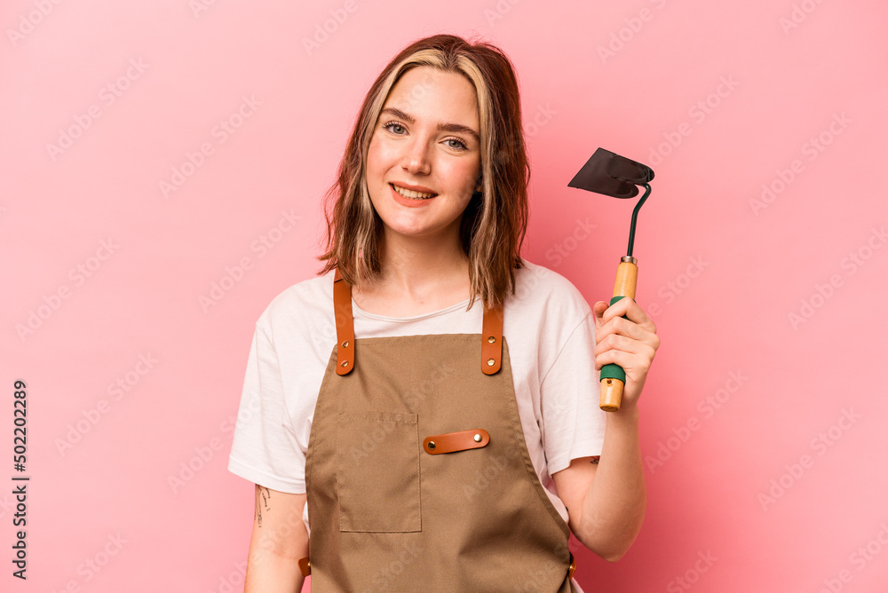 Young gardener woman holding gardening shovel isolated on pink background happy, smiling and cheerful. Young gardener woman holding gardening shovel isolated on pink background happy, smiling and cheerful.