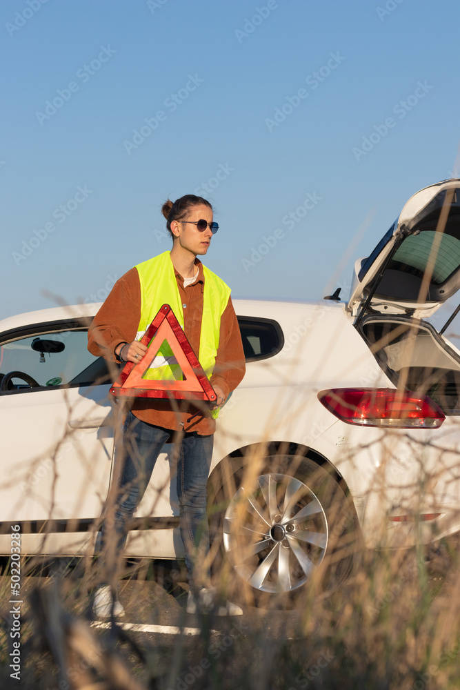Young man on yellow vest setting a road side warning red triangle, warning oncoming traffic of a broken down car