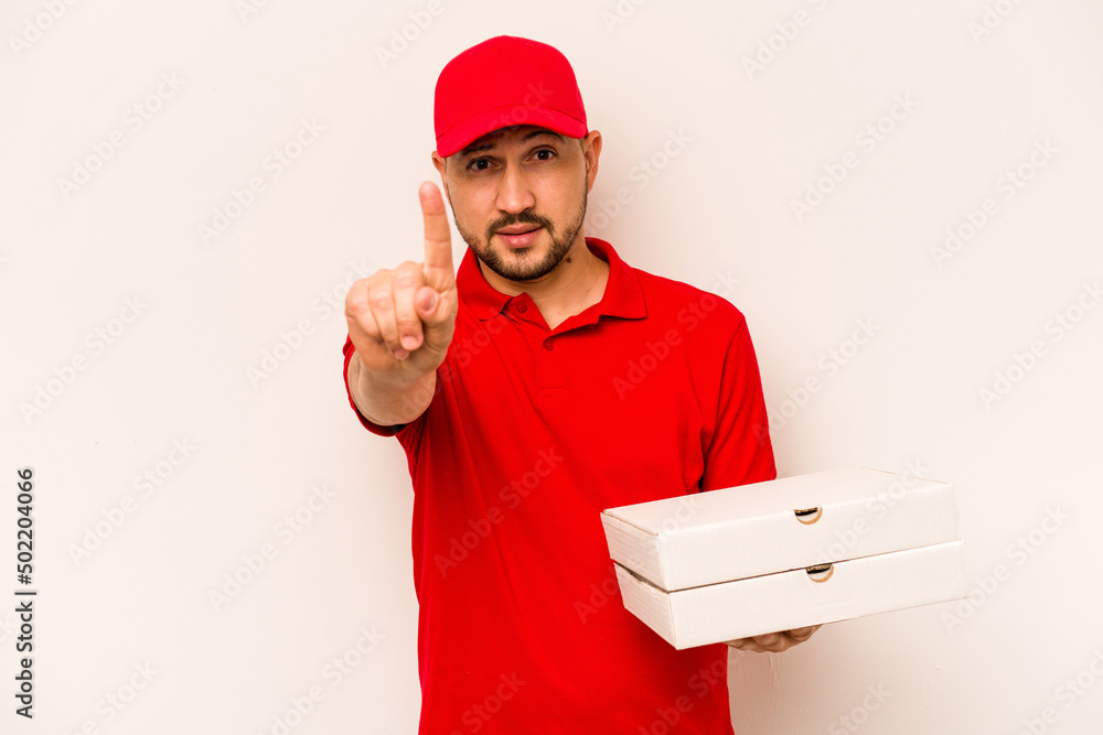 Young delivery man holding pizzas isolated on beige background showing number one with finger. Young delivery man holding pizzas isolated on beige background showing number one with finger.