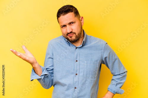 Young hispanic man isolated on yellow background doubting and shrugging shoulders in questioning gesture.