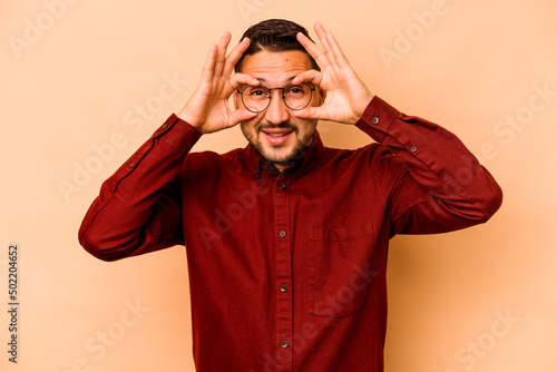 Young hispanic man isolated on beige background showing okay sign over eyes