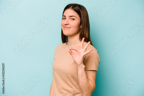 Young caucasian woman isolated on blue background winks an eye and holds an okay gesture with hand.