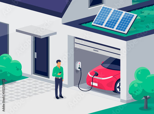 Electric car parking charging inside home garage and green roof wall box charger station. Man standing with smartphone near family house building with clean renewable energy photovoltaic solar panels.