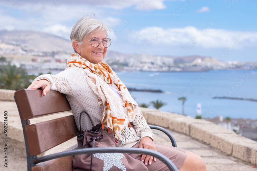 Fototapeta premium Smiling beautiful senior woman with eyeglasses sitting outdoors on bench looking at sea enjoying freedom and vacation. Active elderly lady relaxing on a sunny day