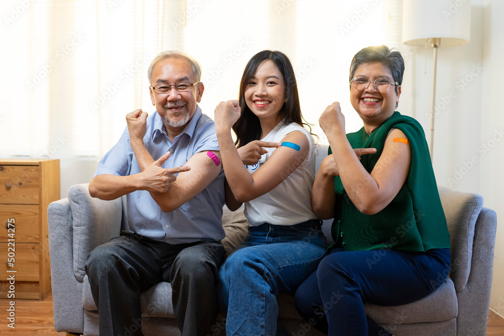 Senior Asian couple and young woman get vaccinated with bandage on arm show thumb up sign in living room. Grandfather and grandmother get vaccine. Senior Asian couple and young woman get vaccinated with bandage on arm show thumb up sign in living room. Grandfather and grandmother get vaccine.