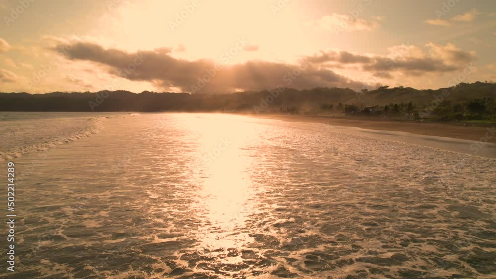 AERIAL: Flying over shallow water at sunset with sun glare reflection. Gentle touches of golden light on an empty tropical beach in Panama. Cloud covered sun reflecting in ocean water. AERIAL: Flying over shallow water at sunset with sun glare reflection. Gentle touches of golden light on an empty tropical beach in Panama. Cloud covered sun reflecting in ocean water.