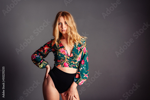 Young blonde woman leaning on one hip in a blouse with a floral print on a gray background