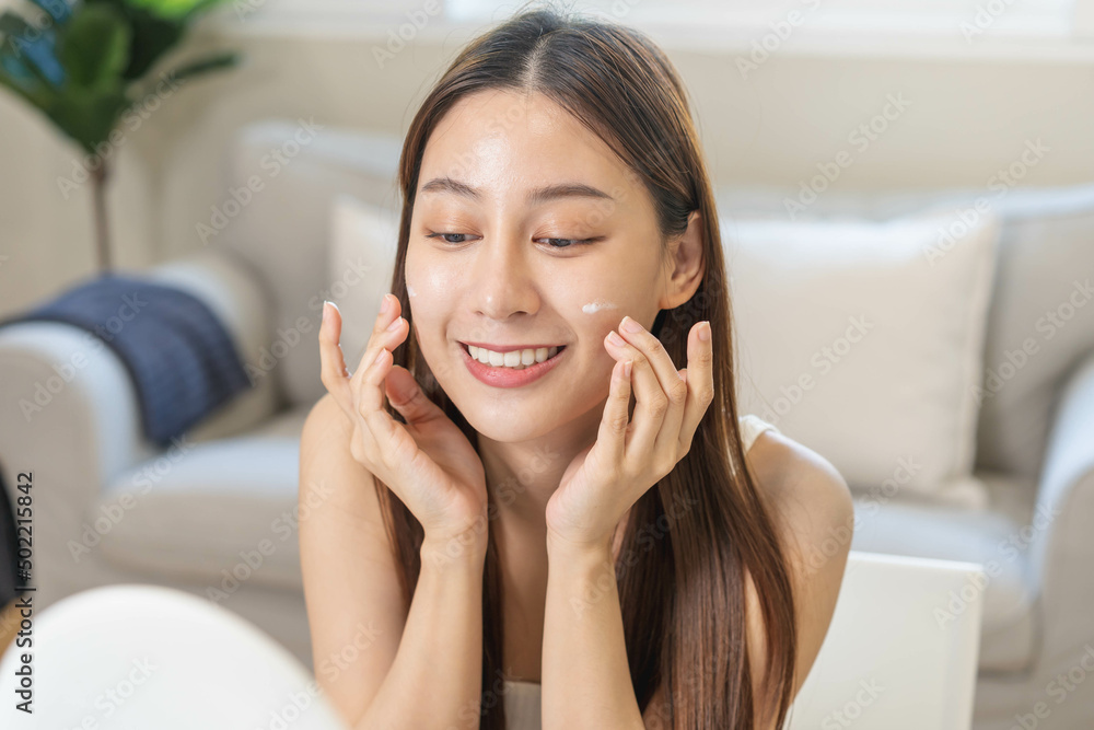 Fresh healthy skin, beautiful smile of asian young woman, girl looking at mirror, applying moisturizer on her face, putting cream treatment before makeup cosmetic routine at home. Facial Beauty. Fresh healthy skin, beautiful smile of asian young woman, girl looking at mirror, applying moisturizer on her face, putting cream treatment before makeup cosmetic routine at home. Facial Beauty.