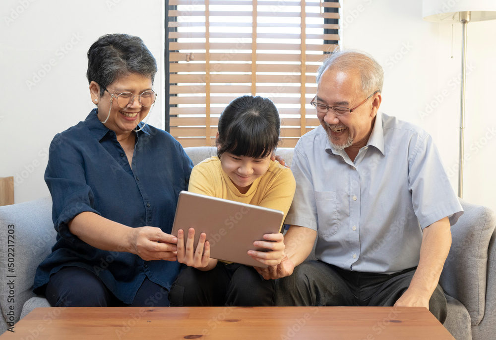 Smiling Asian grandparents on couch with granddaughter looking at tablet. happy three generation family spending time together at home. Smiling Asian grandparents on couch with granddaughter looking at tablet. happy three generation family spending time together at home.