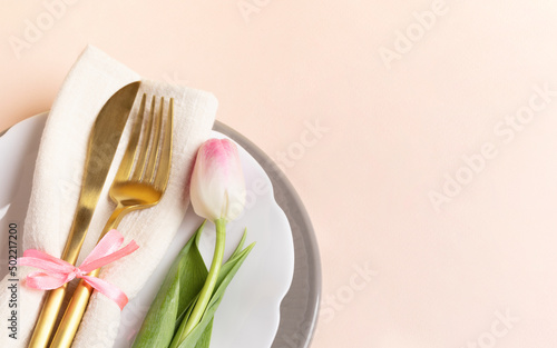 Romantic table setting with golden cutlery and one pink tulip on pastel pink. Copy space. Close up.