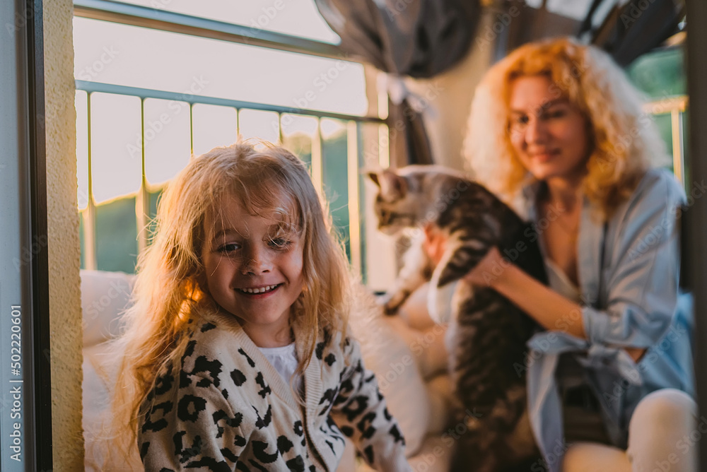 a little, happy girl, a child with a smile plays on a summer, spring vernada at home with mom and a cat at sunset. family weekend evening. cat, pets as a family member. a little, happy girl, a child with a smile plays on a summer, spring vernada at home with mom and a cat at sunset. family weekend evening. cat, pets as a family member.