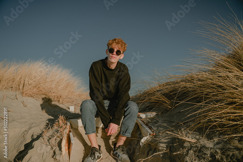 a guy in round glasses and a black jacket sits on the sand dunes