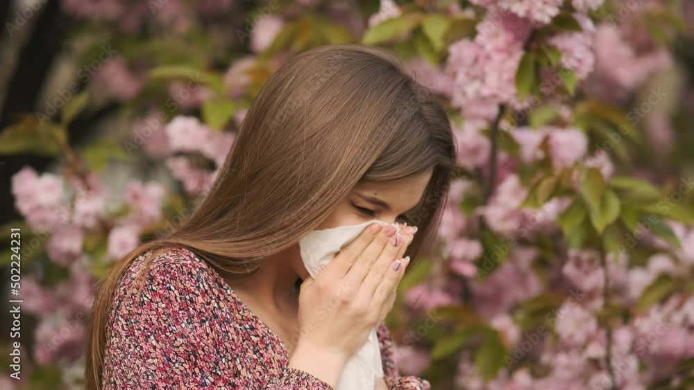 Allergy, sneezing, spring. Woman sneezing in front of blooming tree. Spring allergy concept. Sneezing young girl with nose wiper among blooming trees in park. Pollen allergy, girl sneezing