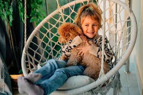 happy little girl, child hugging with a smile her pet, poodle dog at home on the balcony in spring, summer in a cotton-fringed hammock chair at sunset.