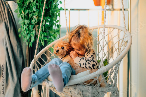 happy little girl, child hugging with a smile her pet, poodle dog at home on the balcony in spring, summer in a cotton-fringed hammock chair at sunset.