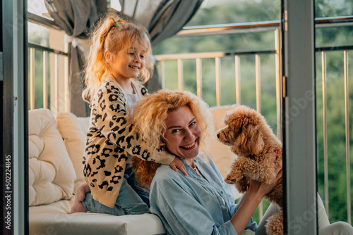 a little, happy girl, a child with a smile plays on a summer, spring vernada at home with mom and a dog at sunset. family weekend evening. dog, pets as a family member.