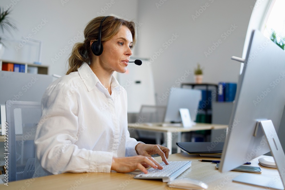Female call center operator wearing microphone headset sitting at the computer in office Female call center operator wearing microphone headset sitting at the computer in office