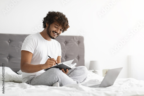 Dark-skinned guy in pajamas using laptop, taking notes