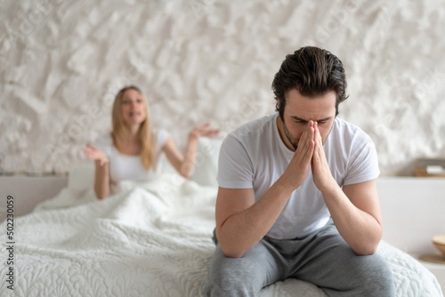 Young man feeling depressed, sitting on bed at home, his angry wife shouting at him, free space. Family problems concept photo