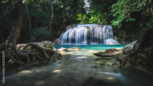 Scenic view of breathtaking waterfall smooth flowing water and emerald green pond in lush rainforest with tree root foreground. Erawan Waterfall, Kanchanaburi, Thailand. Long exposure. © Chavakorn