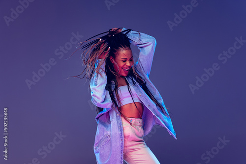 Emotional african girl in jeans and shirt dancing  having fun isolated on dark blue background. Concept of beauty  art  fashion  youth and emotions