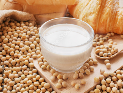 Hot cup of soy milk on lots of soy beans on wooden cutting board with sack of soy beans with some croissants and breads
