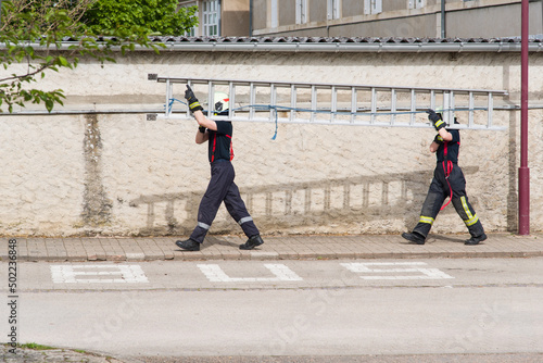 Fotografie Deux jeunes pompiers portant une échelle dans une rue devant un arrêt de bus
